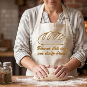 Może przedstawiać: Beżowy fartuch ze złotym obrysem bochenka chleba i napisem "Give us this day our daily bread". Osoba ugniata ciasto na drewnianej powierzchni. Słoiki z przyprawami i miedziana miska w tle.