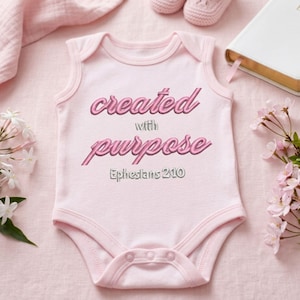 Puede incluir: Un body de bebé rosa claro con las palabras "created with purpose" y "Ephesians 2:10" en escritura rosa. La imagen incluye zapatos de bebé, una biblia y flores.