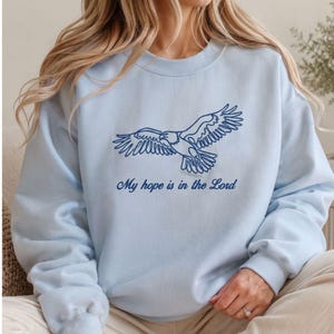 May include: A light blue sweatshirt featuring a detailed navy blue outline of an eagle in flight. Below the eagle is the text "My hope is in the Lord". The sweatshirt is a comfortable, casual style.