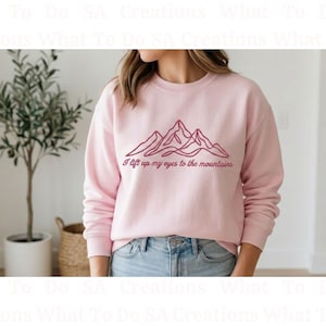 Może przedstawiać: Jasnoróżowa bluza z napisem "I lift up my eyes to the mountains" i grafiką gór w ciemniejszym różu. Bluza z długim rękawem jest połączona z niebieskimi dżinsami.