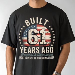 May include: Black t-shirt with a distressed graphic. The graphic features the text "BUILT 61 YEARS AGO" in a circular design with an American flag background. Additional text reads "Original & Unrestored" and "MOST PARTS STILL IN WORKING ORDER."
