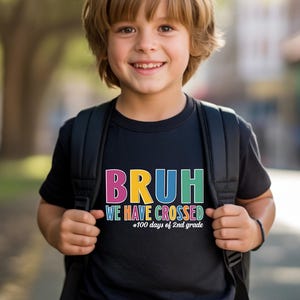 May include: A black short-sleeved t-shirt featuring the colourful text BRUH WE HAVE CROSSED 100 days of 2nd grade printed on the front. The shirt is worn with a black backpack, showcasing a casual style with vibrant lettering on a dark background.