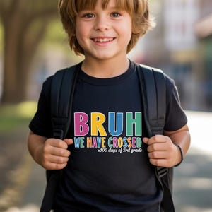 May include: A black short-sleeved t-shirt featuring the colourful text BRUH WE HAVE CROSSED 100 days of 3rd grade printed on the front. The shirt is worn with a black backpack, and the design uses pink, yellow, blue, and green lettering.