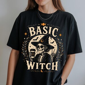 May include: A black short-sleeved t-shirt featuring a graphic print of a skeleton wearing a witch hat, holding a coffee cup, and making a peace sign. The text BASIC WITCH is printed in a cream-colored font, surrounded by stars, leaves, and moons.