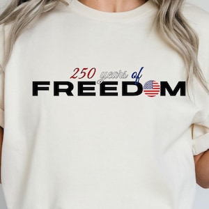 Puede incluir: Camiseta color crema con el texto "250 years of FREEDOM" en negro, rojo y azul. La letra "O" en "FREEDOM" se reemplaza con un diseño de la bandera estadounidense. La camiseta es de estilo casual.