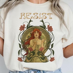 Op de afbeelding: Een crèmekleurig t-shirt met het woord RESIST in een gestileerd lettertype boven een grafische illustratie van een persoon met lang rood haar, gekleed in een lichtgekleurd kledingstuk, omlijst door groene ranken en rode bloemen.