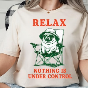 Könnte beinhalten: Cremefarbenes T-Shirt mit den orangefarbenen Worten "RELAX" und "NOTHING IS UNDER CONTROL". Ein Cartoon-Mops mit Sonnenbrille und Hut sitzt in einem grünen Klappstuhl.