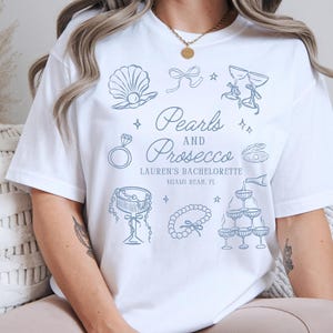 Può includere: T-shirt bianco con un design grafico azzurro che presenta una conchiglia, un anello, un fiocco, calici di champagne e il testo "Pearls and Prosecco". Il testo include anche "Lauren's Bachelorette" e "Miami Beach, FL".
