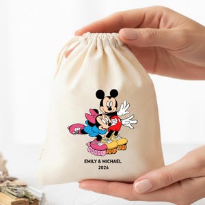 May include: A cream-colored drawstring bag featuring a cartoon image of Mickey and Minnie Mouse on roller skates. The image includes the text "EMILY & MICHAEL 2026" in black. The bag is held by two hands.