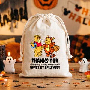 Puede incluir: Una bolsa de lona color crema con cordón que presenta un gráfico de Winnie the Pooh y Tigger con disfraces de Halloween. El texto dice: THANKS FOR Visiting My Spooky Hunny Patch! NOAH'S 1ST HALLOWEEN. La bolsa tiene dos cuerdas blancas.