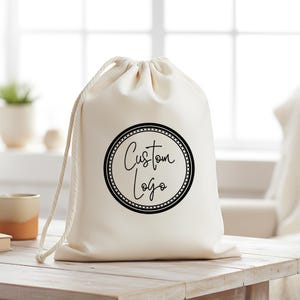 May include: Cream-coloured drawstring bag featuring a black circular logo with the words "Custom Logo" in cursive. The bag is made of a soft fabric and has a white drawstring closure. It rests on a wooden surface.