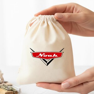 May include: A natural-colored cotton drawstring pouch featuring a red and black graphic emblem on the front. The emblem is shaped like a stylized car hood ornament with the name Noah written in white script across the center of the red section.