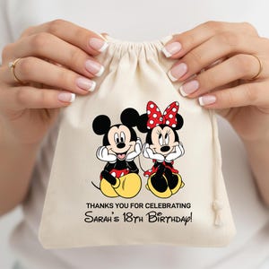 May include: A beige drawstring bag featuring Mickey and Minnie Mouse. Mickey is wearing red shorts and yellow shoes, while Minnie wears a red polka dot bow and dress. The text reads, "THANKS YOU FOR CELEBRATING Sarah's 18th Birthday!"