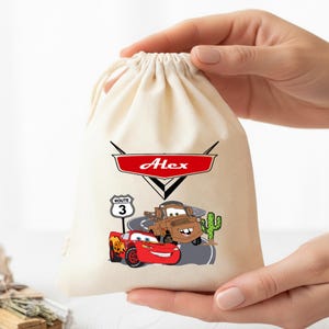 Puede incluir: Una bolsa de color crema con cord&oacute;n que presenta un emblema gr&aacute;fico rojo con el nombre Alex impreso en blanco. Debajo, un coche de carreras rojo y una gr&uacute;a marr&oacute;n aparecen sobre una carretera junto a una se&ntilde;al de Ruta 3 y un cactus verde.