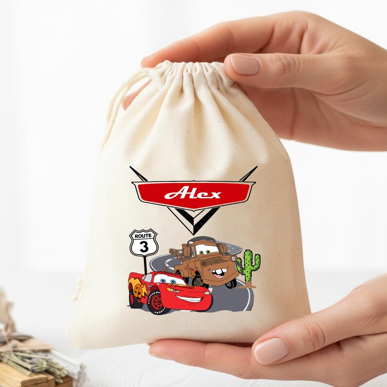 Puede incluir: Una bolsa de color crema con cord&oacute;n que presenta un emblema gr&aacute;fico rojo con el nombre Alex impreso en blanco. Debajo, un coche de carreras rojo y una gr&uacute;a marr&oacute;n aparecen sobre una carretera junto a una se&ntilde;al de Ruta 3 y un cactus verde.