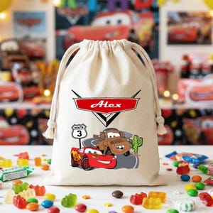 Puede incluir: Una bolsa de algod&oacute;n beige con cord&oacute;n que presenta un gr&aacute;fico rojo y blanco con el nombre Alex dentro de un logotipo de coche, con ilustraciones de un coche de carreras rojo, una gr&uacute;a marr&oacute;n, una se&ntilde;al de Ruta 3 y un cactus verde.