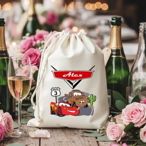 Puede incluir: Una bolsa de color crema con cord&oacute;n que presenta un estampado gr&aacute;fico de un coche de carreras rojo y una gr&uacute;a marr&oacute;n en una carretera con un cactus. Sobre los veh&iacute;culos hay un emblema rojo con el nombre Alex y una se&ntilde;al de Ruta 3.