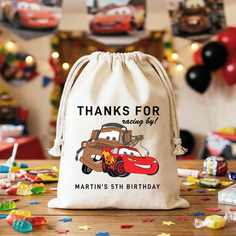 May include: A beige drawstring bag featuring a cartoon image of two Cars characters, Lightning McQueen and Mater. The bag has the text "THANKS FOR racing by!" and "MARTIN'S 5TH BIRTHDAY" printed on it. The bag is sitting on a wooden surface.