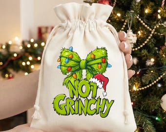Könnte beinhalten: Ein cremefarbener Geschenkbeutel mit Kordelzug und einem grünen Schleifendesign, das mit bunten Weihnachtslichtern verziert ist. Der Beutel trägt den Text "NOT GRINCHY" in Grün und Gelb und einen kleinen Weihnachtsmannhut.