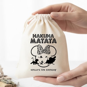 May include: A cream-colored drawstring bag with the words "HAKUNA MATATA" in bold black letters. Below the text is a black outline of Mickey Mouse ears with a leopard print bow. The bag also features silhouettes of Simba, Timon, and Pumbaa, with the text "Stella's 5th Birthday".