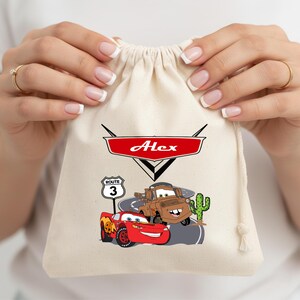 Puede incluir: Una bolsa de color crema con cord&oacute;n que presenta un emblema gr&aacute;fico rojo con el nombre Alex en letras blancas. Debajo, un coche de carreras rojo y una gr&uacute;a marr&oacute;n aparecen sobre una carretera junto a una se&ntilde;al de Ruta 3 y un cactus verde.