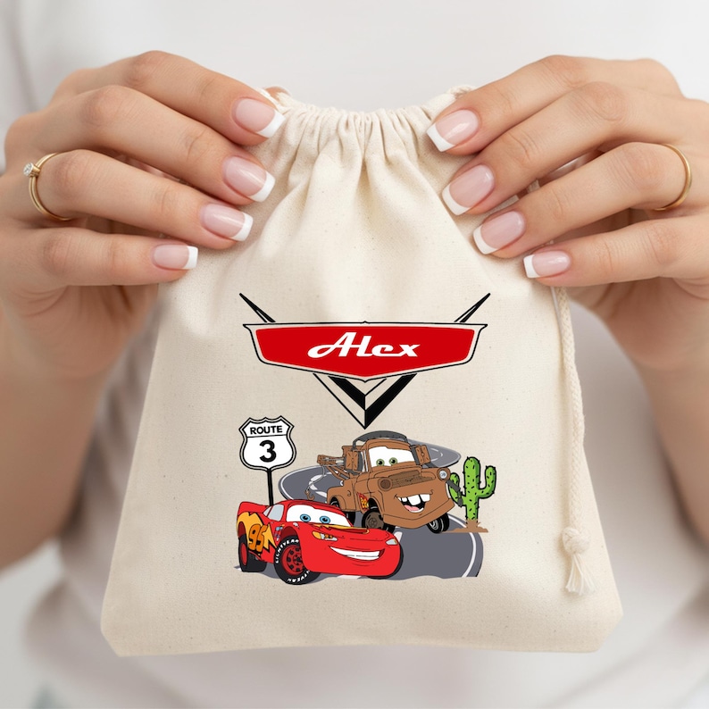 Puede incluir: Una bolsa de color crema con cord&oacute;n que presenta un emblema gr&aacute;fico rojo con el nombre Alex en letras blancas. Debajo, un coche de carreras rojo y una gr&uacute;a marr&oacute;n aparecen sobre una carretera junto a una se&ntilde;al de Ruta 3 y un cactus verde.