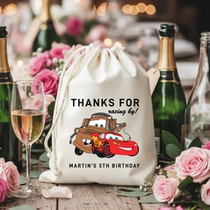 May include: A white drawstring bag with the text "THANKS FOR racing by!" and "MARTIN'S 5TH BIRTHDAY". The bag features a cartoon illustration of two cars. The background includes champagne bottles, glasses, and pink roses.