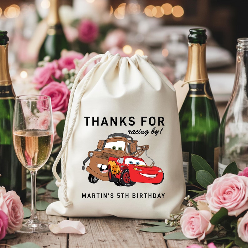 May include: A white drawstring bag with the text "THANKS FOR racing by!" and "MARTIN'S 5TH BIRTHDAY". The bag features a cartoon illustration of two cars. The background includes champagne bottles, glasses, and pink roses.