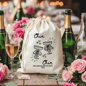 Puede incluir: Una bolsa de cordón de color crema con un gráfico negro de dos copas de champán y la palabra "Chin" arriba y abajo. El texto "MIA'S BACHELORETTE PARTY 2025" está impreso debajo. El fondo incluye botellas de champán y rosas rosas.