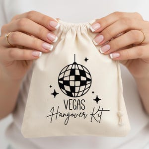May include: A beige drawstring bag with a black disco ball graphic and the words "VEGAS Hangover Kit". The bag is held by hands with manicured nails and gold rings. The design includes four star shapes.