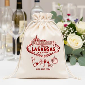 Puede incluir: Una bolsa de cordón de color crema con un gráfico rojo del letrero de Las Vegas, champán y una bola de discoteca. Se muestra el texto "WELCOME to Fabulous LAS VEGAS NEVADA". Debajo del gráfico está el texto "GIRL TRIP 2026".