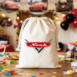 May include: A natural-colored canvas drawstring bag featuring a red and black graphic emblem on the front. The emblem is shaped like a stylized car hood ornament with the name Noah printed in white script across the center of the red section.