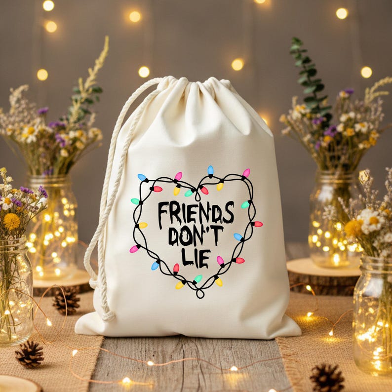 Może przedstawiać: Kremowy worek ze sznurkiem z napisem "FRIENDS DON'T LIE" wewnątrz serca z kolorowych lampek choinkowych. Worek wykonany jest z miękkiej tkaniny i ma białe zamknięcie ze sznurkiem.