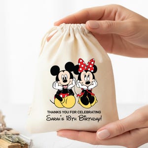 May include: A cream-colored drawstring bag featuring Mickey and Minnie Mouse illustrations. Mickey wears red shorts, and Minnie has a red polka-dot bow. Text reads, "THANKS YOU FOR CELEBRATING Sarah's 18th BIRTHDAY!"
