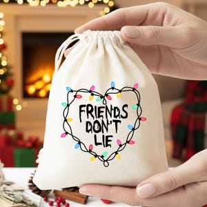 May include: A cream-coloured drawstring bag featuring the text "FRIENDS DON'T LIE" within a heart-shaped string of colourful fairy lights. The bag is held against a blurred background.