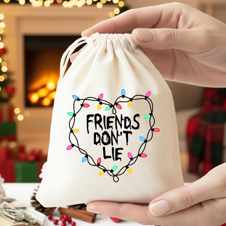 Może przedstawiać: Kremowy worek ze sznurkiem z napisem "FRIENDS DON'T LIE" wewnątrz serca z kolorowych lampek choinkowych. Worek jest trzymany na tle rozmytego tła.
