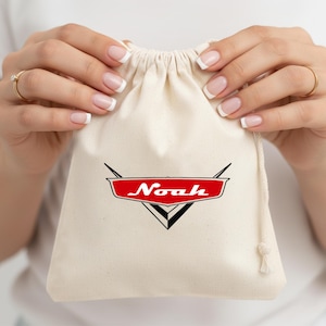 May include: A cream-colored fabric drawstring pouch featuring a red and black graphic emblem on the front. The emblem is shaped like a stylized car logo with the name Noah printed in white script lettering across the center of the red section.