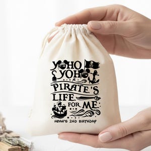 Puede incluir: Una bolsa de cordón de color crema con un diseño de pirata negro. El texto dice "Yo Ho Yo Ho, A Pirate's Life For Me" con un barco pirata, calavera y huesos cruzados, y un ancla. La bolsa también dice "Noah's 2nd Birthday".