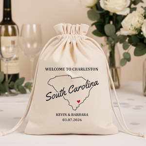 May include: A beige drawstring bag with the text "WELCOME TO CHARLESTON" above a South Carolina outline and the words "South Carolina" in cursive. Below is "KEVIN & BARBARA" and the date "03.07.2026". A red heart is inside the state outline.