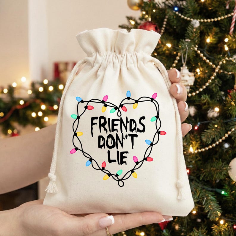 Może przedstawiać: Kremowy worek na prezent ze sznurkiem z napisem "FRIENDS DON'T LIE" wewnątrz serca z kolorowych lampek choinkowych. Torba jest trzymana przed choinką.