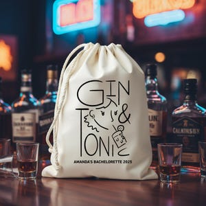 May include: Cream-colored drawstring bag with the words "GIN & TONIC" in black, along with a graphic of a person and a cocktail. The text "AMANDA'S BACHELORETTE 2025" is printed below. The bag is on a wooden surface with liquor bottles and shot glasses in the background.