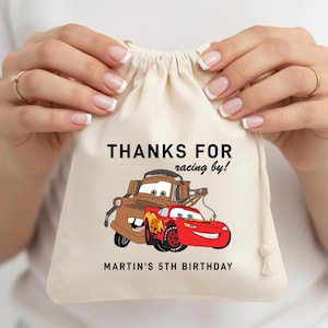 May include: A beige drawstring bag featuring the text "THANKS FOR racing by!" and "MARTIN'S 5TH BIRTHDAY". The bag has a cartoon illustration of two Cars characters, Tow Mater and Lightning McQueen.