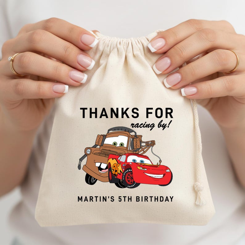May include: A beige drawstring bag featuring the text "THANKS FOR racing by!" and "MARTIN'S 5TH BIRTHDAY". The bag has a cartoon illustration of two Cars characters, Tow Mater and Lightning McQueen.
