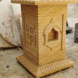 May include: A tan sandstone pedestal featuring a carved lotus flower top, a square base with decorative borders, a swastika symbol on one side, and an arched niche on the front panel. The structure is crafted with detailed geometric patterns.