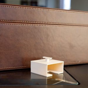 May include: A brown leather box with stitched detailing, a white rectangular object with a handle, and a glass of liquid on a dark surface. The box is rectangular and has a textured surface.