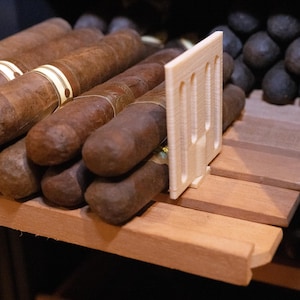 May include: A collection of brown cigars with paper bands arranged on a wooden shelf, separated by a vertical, off-white, rectangular plastic divider with three vertical slots, positioned next to a pile of dark, rounded objects.