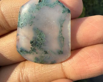 May include: A polished gemstone with a unique pattern. The stone features a pale, translucent base with intricate green and black inclusions, creating a natural, artistic design. The shape is a modified hexagon with rounded edges.