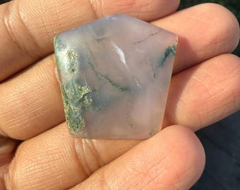 May include: A translucent, irregularly shaped gemstone with a pale lavender hue. The stone features internal patterns of white and green, with a mossy green patch on one edge. The stone is held in a hand.