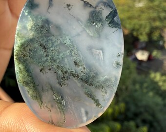 May include: Oval-shaped gemstone with a translucent, milky white base. The stone features intricate patterns of green moss-like inclusions, creating a natural, artistic design. The gemstone is held between fingers.