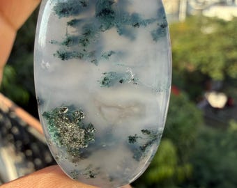 May include: An oval-shaped gemstone with a translucent white base, featuring intricate patterns of dark green and gray inclusions. The polished surface reflects light, highlighting the natural textures and colors of the stone.